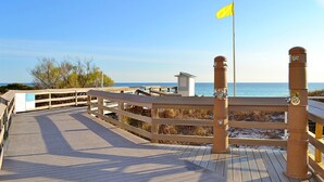 Un paseo marítimo de madera que lleva a una playa con un asta de banderas y una pequeña estructura.