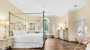 Un dormitorio con cama con dosel, ventilador de techo, suelo de madera, cómoda y ventana con persianas.
