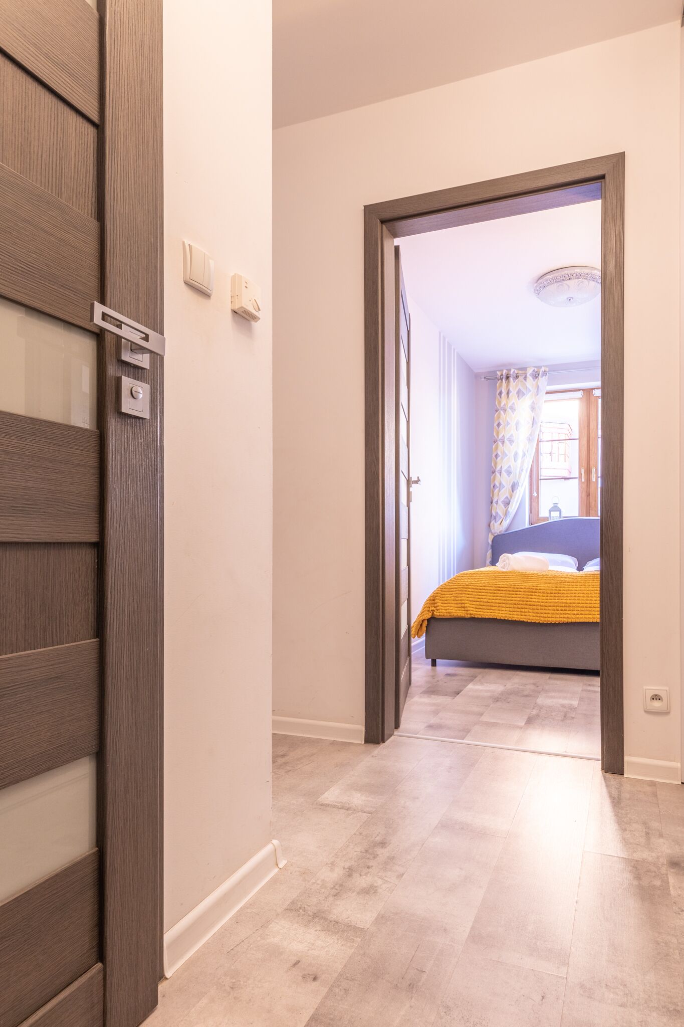 Een gang die leidt naar een slaapkamer met een bed, een deur en een lichtschakelaar aan de muur.