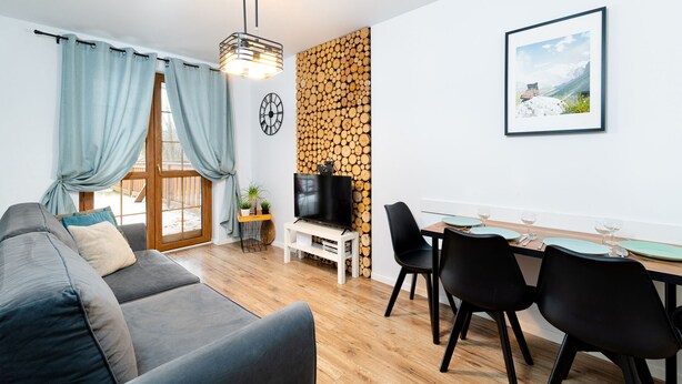Una sala de estar moderna con un sofá gris, una mesa de comedor con sillas, un televisor sobre un mueble bajo y un reloj en la pared.