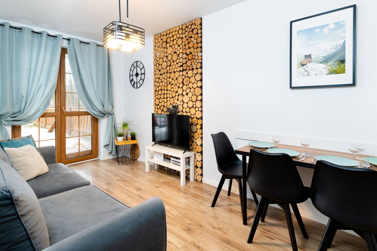 A modern living room with a grey sofa, a dining table with chairs, a TV on a stand, and a wall-mounted clock.
