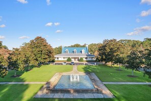 Stately mansion with a central entrance and manicured lawns, surrounded by mature trees and a serene water feature.