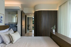 A modern bedroom with a large bed, a wooden headboard, a bedside table with a plant, and a view of a bathroom and a hallway.