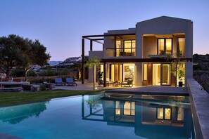 A modern two-story house with a swimming pool, outdoor seating, and a well-lit interior.