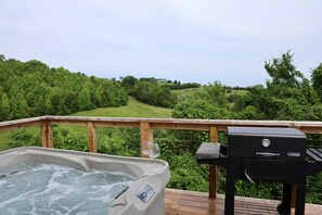 A hot tub and a grill on a wooden deck with a view of a lush green landscape.