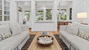 A spacious living room with white furniture, a wooden coffee table with seashells, and a view of the kitchen and dining area through open doors. - 5-Bedroom WaterColor Home | Breathtaking Gulf & Lake Views | Beach Steps Away (SANTA ROSA BEACH)