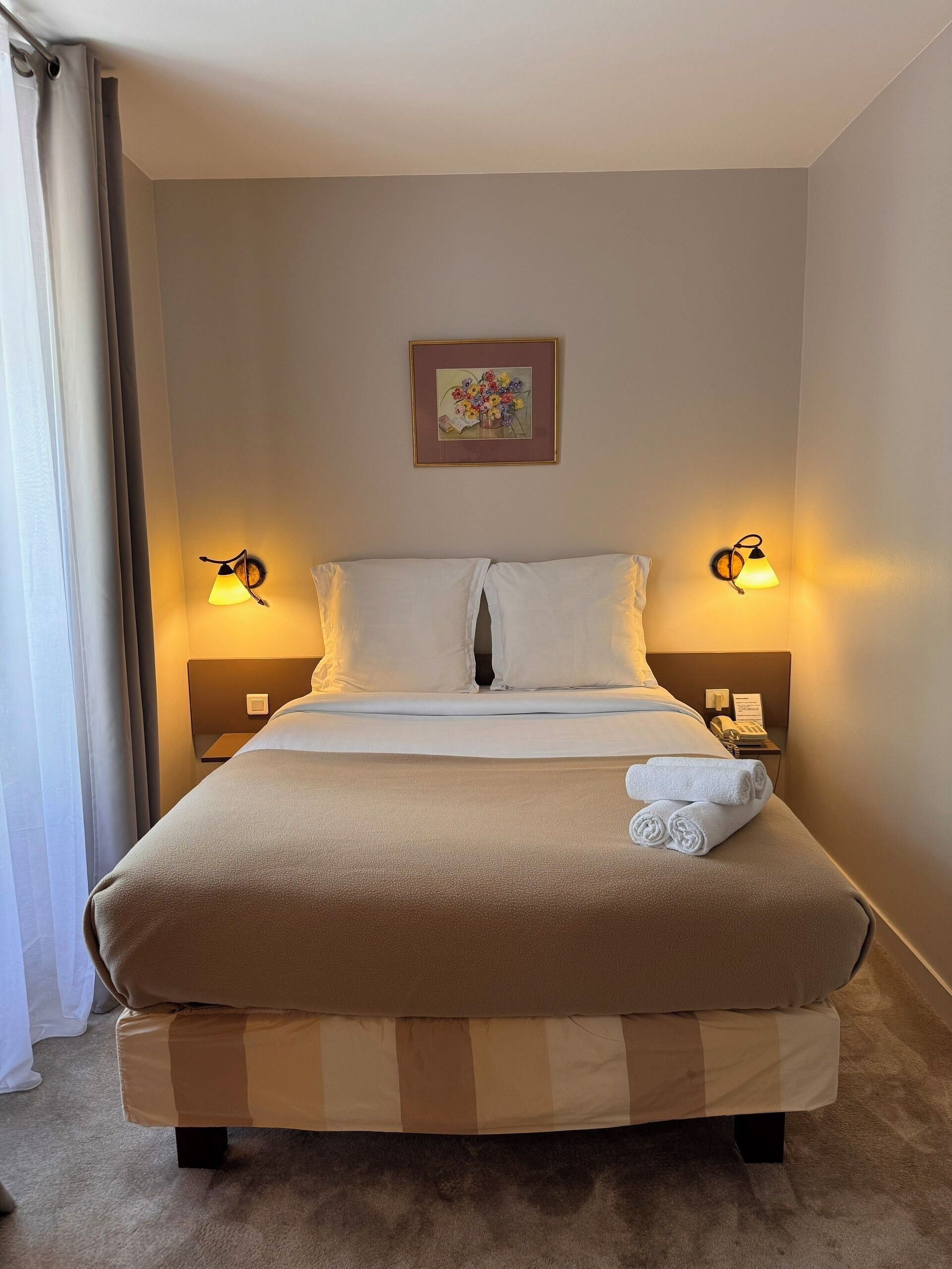 A hotel room with a large bed, two bedside lamps, a framed picture on the wall, and a neatly made bed with a rolled towel.