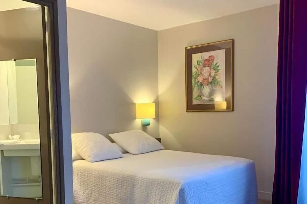 A hotel room with a bed, a nightstand with a lamp, a painting on the wall, and a bathroom visible through an open door.