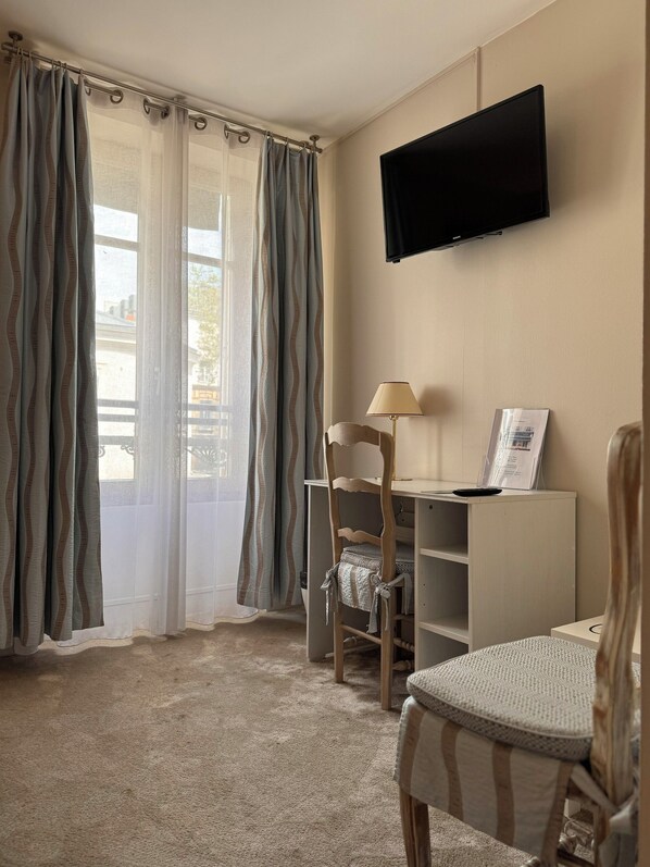 A room with a desk, a chair, a television mounted on the wall, and a window with curtains.