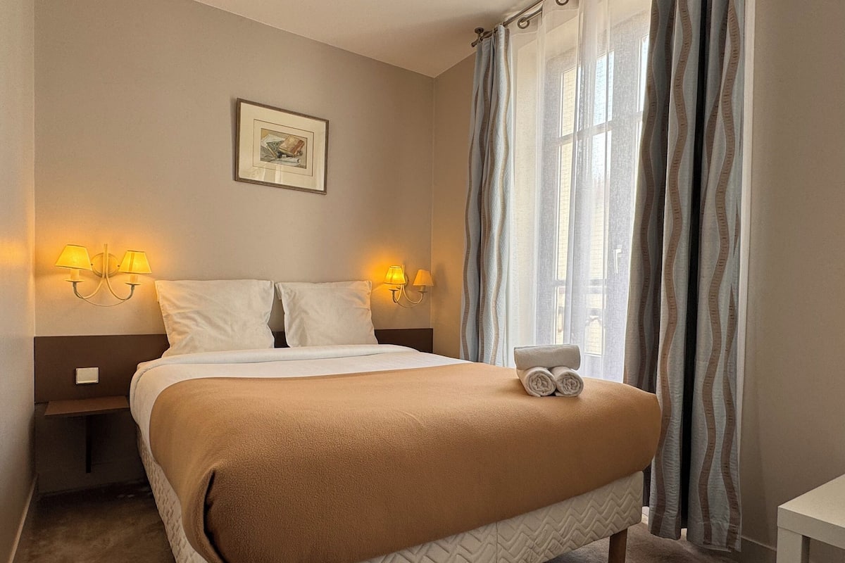 A neatly made bed with a beige blanket, two bedside lamps, a framed picture on the wall, and a window with curtains.