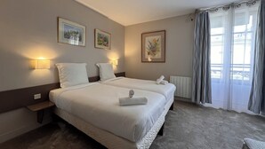 Une chambre d’hôtel avec deux lits, des œuvres d’art encadrées aux murs, une fenêtre avec des rideaux et une table de chevet avec une lampe.