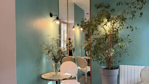 Une pièce aux murs de couleur turquoise, avec une table ronde en bois, deux chaises en osier blanches, une grande plante en pot et un miroir qui reflète la pièce.