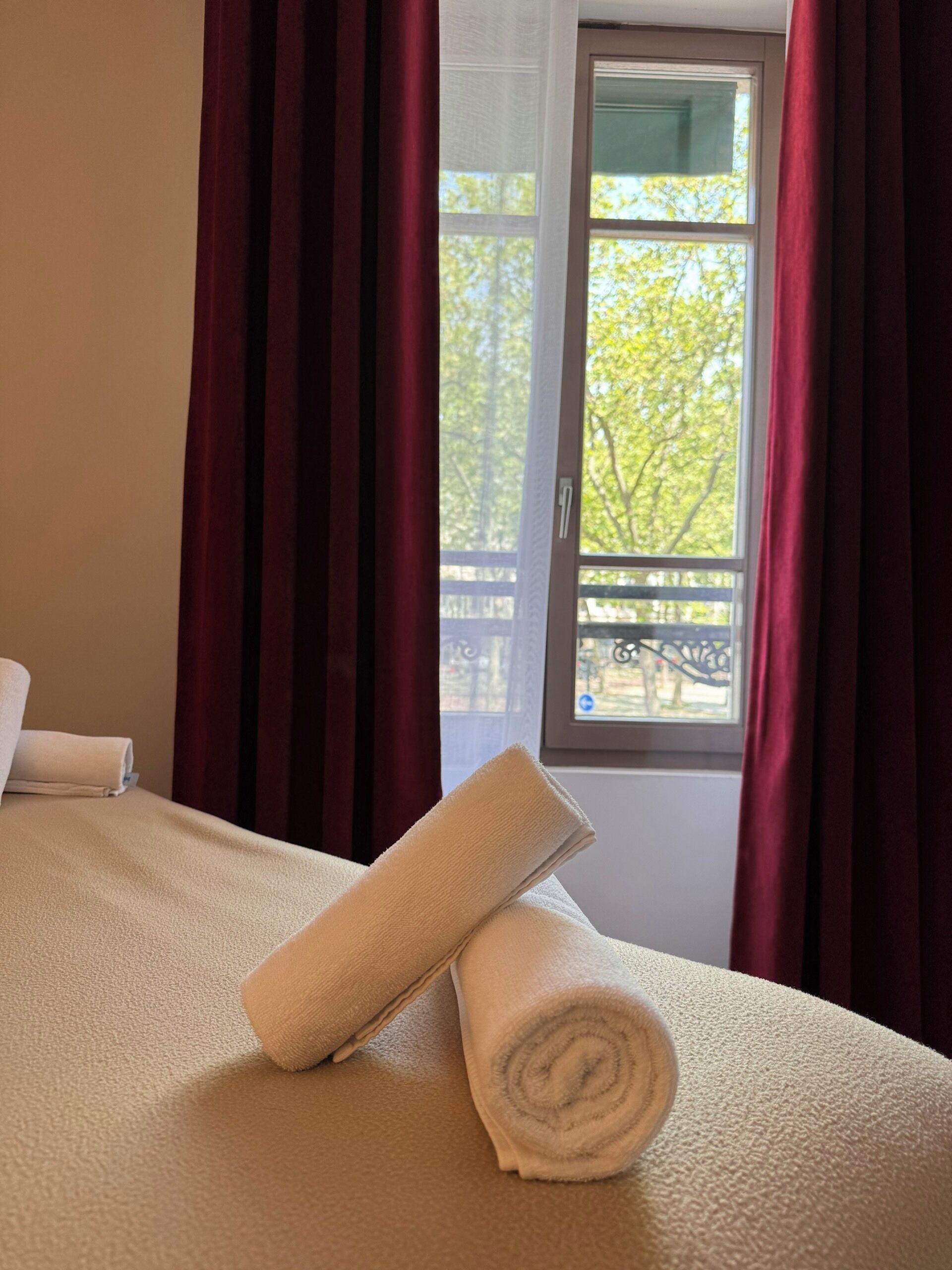 A hotel room with a bed, a window with a view of trees, and red curtains.