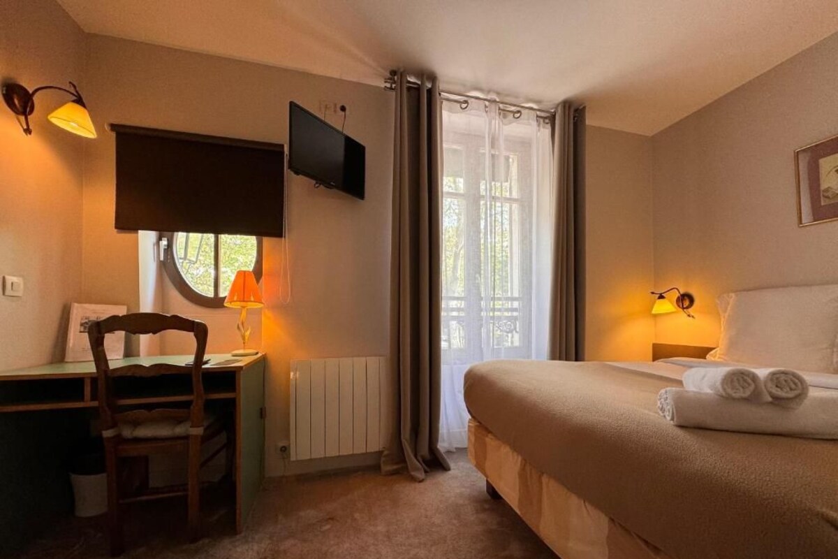 A bedroom with a bed, a desk, a chair, a TV, and a window with curtains.