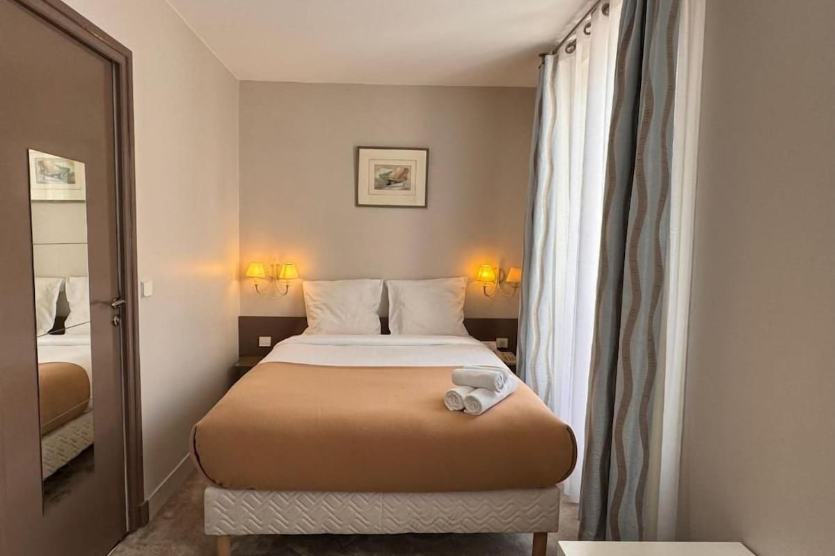 A hotel room with a bed, two bedside lamps, a framed picture on the wall, and a window with curtains.