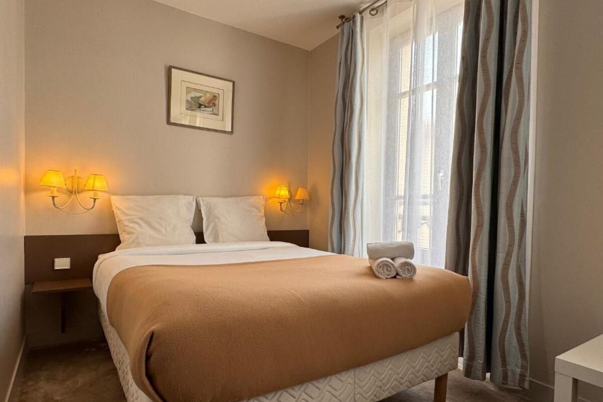 A neatly made bed with a brown blanket, two bedside lamps, a framed picture on the wall, and a window with curtains.
