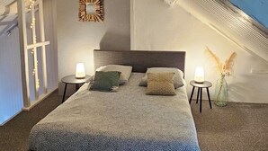 Ett sovrum med ett sluttande tak, en säng med ett mönstrat överkast, två sänglampor, ett litet bord med en lampa och dekorativa växter.