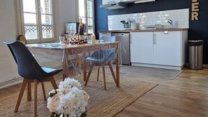 Ett modernt kök med vita skåp och en svart väggbeklädnad, en matplats med ett glasbord och stolar samt ett stort fönster som släpper in naturligt ljus.