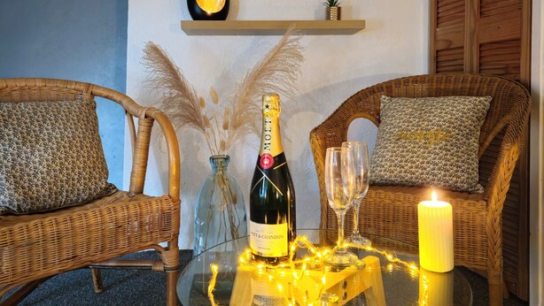En mysig sittgrupp med flätade stolar, ett glasbord med en flaska champagne, två glas och ett tänt ljus.