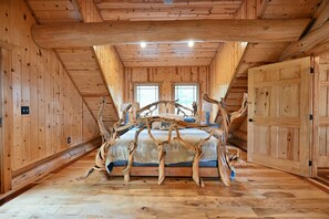 Una habitación con techo inclinado, paredes de madera, una cama con un original cabecero hecho de una rama de árbol y dos ventanas.
