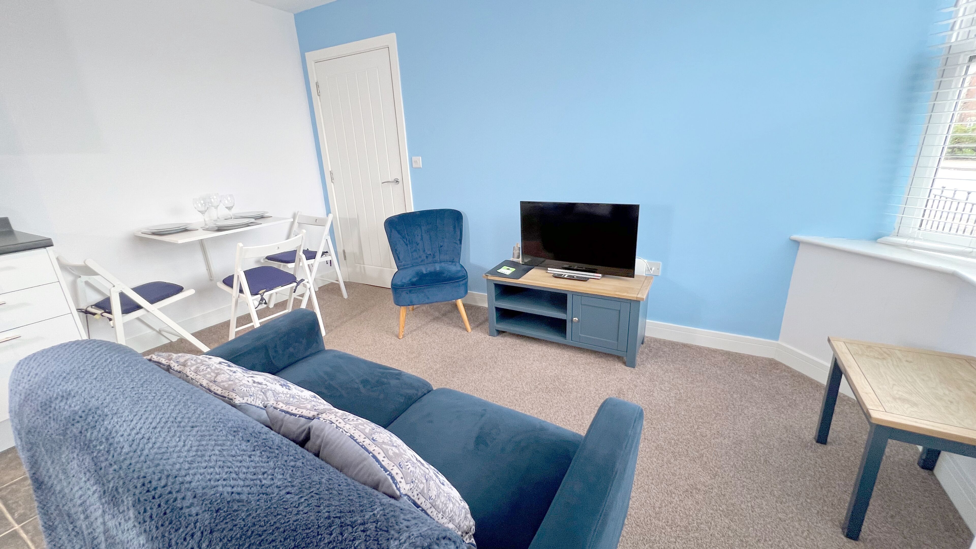 A small, modern living room with a blue sofa, a blue armchair, a TV on a wooden stand, and a small table.