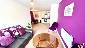 Una sala moderna con un sofá, una mesa de centro y una butaca morada. Se observa una cocina al fondo, con una mesa de comedor y sillas.