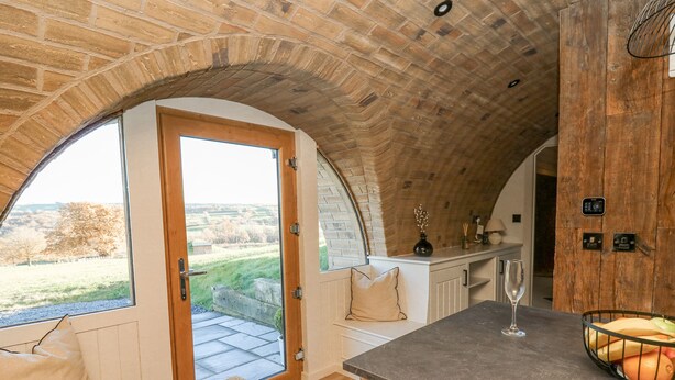 A cozy kitchen with a stone arch ceiling, a wooden bench with cushions, a bar area with stools, and a view of the countryside through a large window. - Tan y Wawr- Onnen (Welshpool)