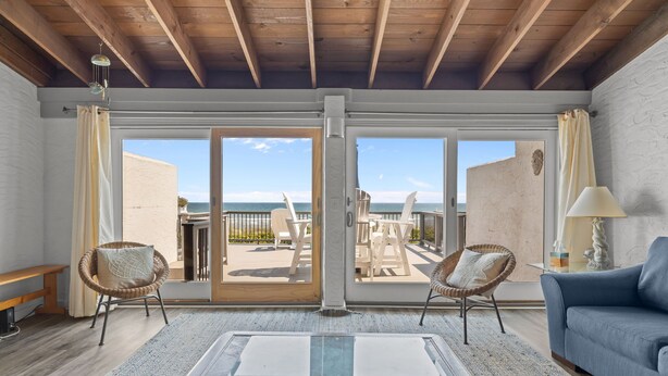 A spacious living room with a large glass table, wicker chairs, a blue sofa, and a view of the beach through a sliding glass door. - 4 BDR-Ocean Front Townhome in Pine Knoll Shores (Oceans 2) (Pine Knoll Shores)