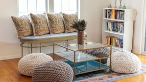 Una sala de estar acogedora con una mesa de centro de vidrio, pufs de punto, una estantería y un asiento junto a la ventana con cojines estampados.
