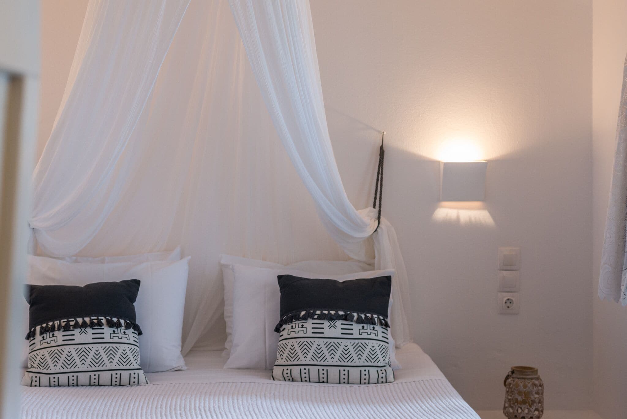 A bed with a canopy, white bedding, patterned pillows, a wall-mounted light, and a small decorative vase.