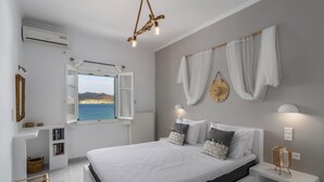 A bedroom with a bed, bedside tables, a window with curtains, a wall-mounted light fixture, and a view of the sea. - Venia's Guesthouse (Milos)