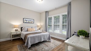 A bedroom with a bed, nightstands, a window with blinds, a rug, and a small table with a plant. - Near downtown Cary & RDU. Easy access to Raleigh, Durham and local venues. (Cary)