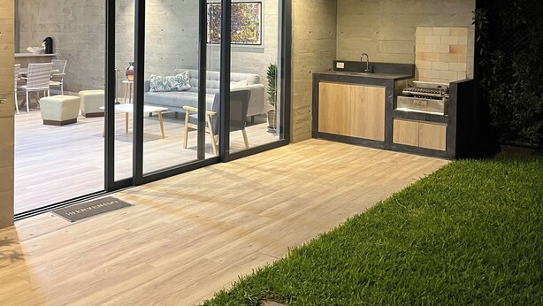 Un espace cuisine extérieur moderne, doté d’un barbecue intégré, d’un coin salon avec un canapé et des fauteuils, et d’une porte vitrée donnant accès à un espace intérieur comprenant une table et des chaises.