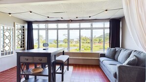 A room with a dining table, a sofa, and large windows with a view of greenery.