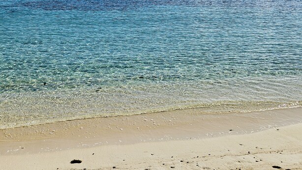 A beach with clear water, pebbles, and distant mountains.