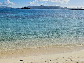 A beach with clear water, pebbles, and distant mountains.