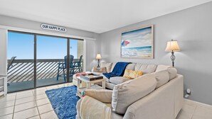 A living room with a sofa, a coffee table, and a blue rug. There is a sliding glass door leading to a balcony with a view of the ocean.