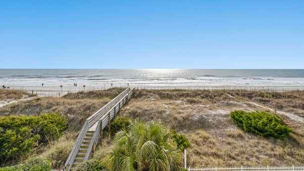 A beach with a wooden boardwalk leading to the water, surrounded by dry grass and sparse vegetation.