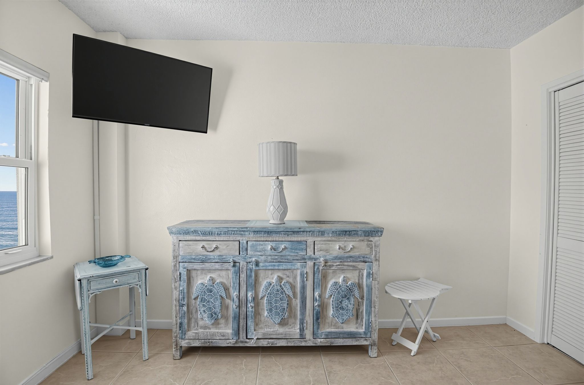 A room with a white cabinet, a small table, a television mounted on the wall, and a window with a view.