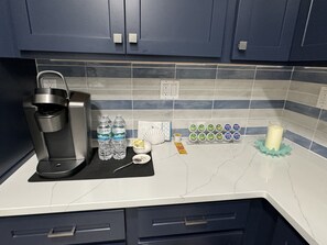 Eine Küche mit dunkelblauen Schränken, einer Kaffeemaschine, Wasserflaschen, einer Zuckerlösung, einem Glas Milch und einer Schale mit Joghurtbechern.
