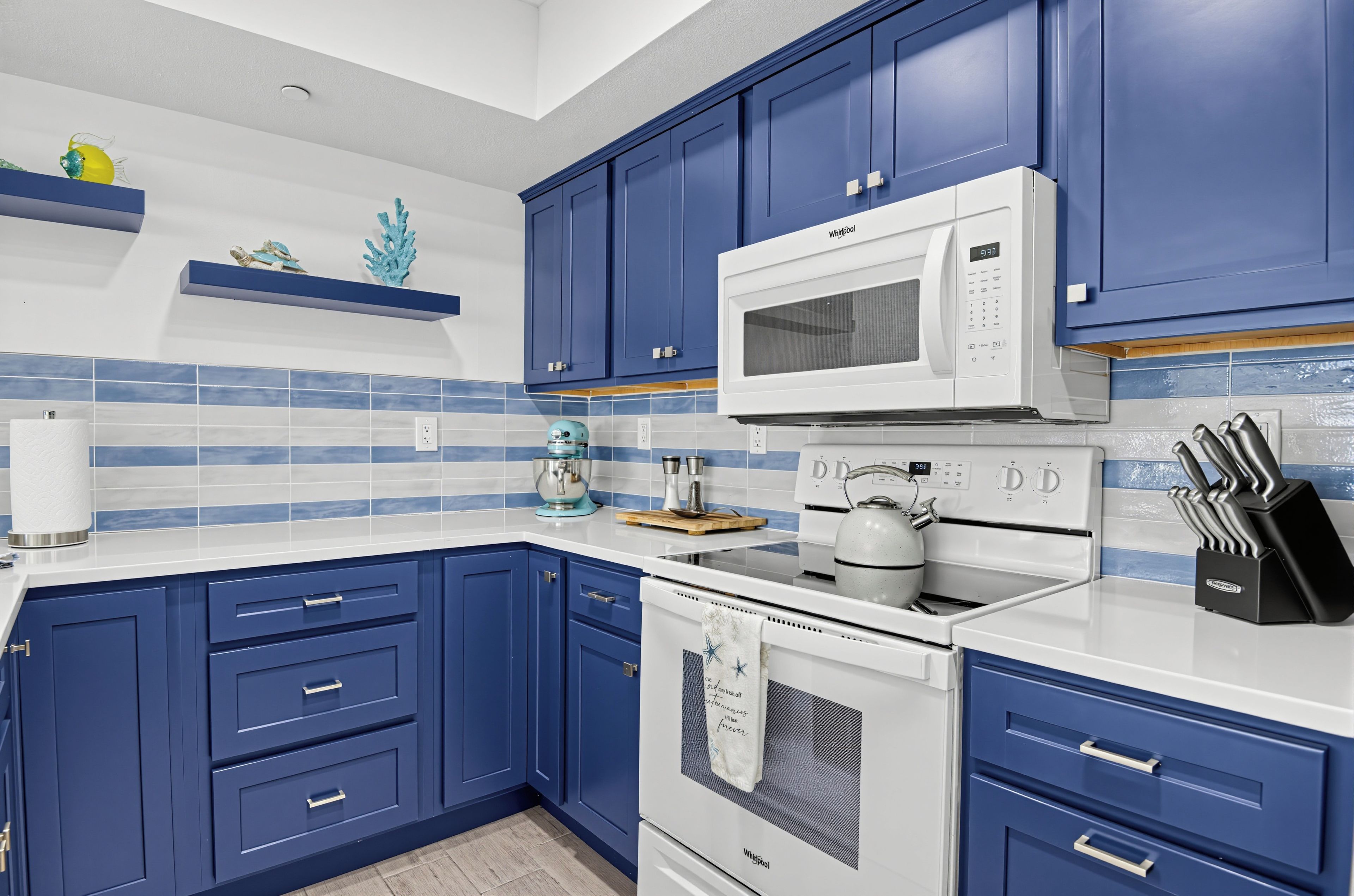 A kitchen with blue cabinets, a white microwave, a kettle, and a knife block.