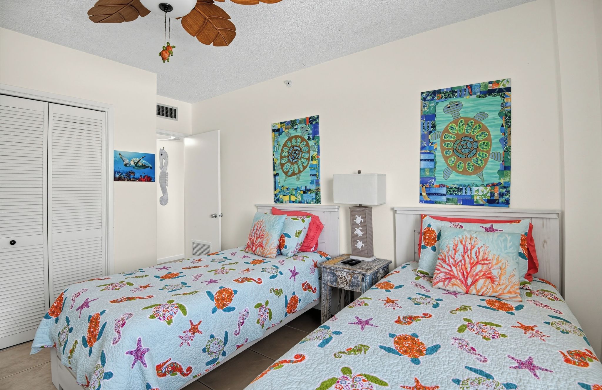 A room with two beds, each with a colorful sea-themed bedspread, a ceiling fan, and artwork on the walls.