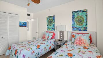 A room with two beds, each with a colorful sea-themed bedspread, a ceiling fan, and artwork on the walls.