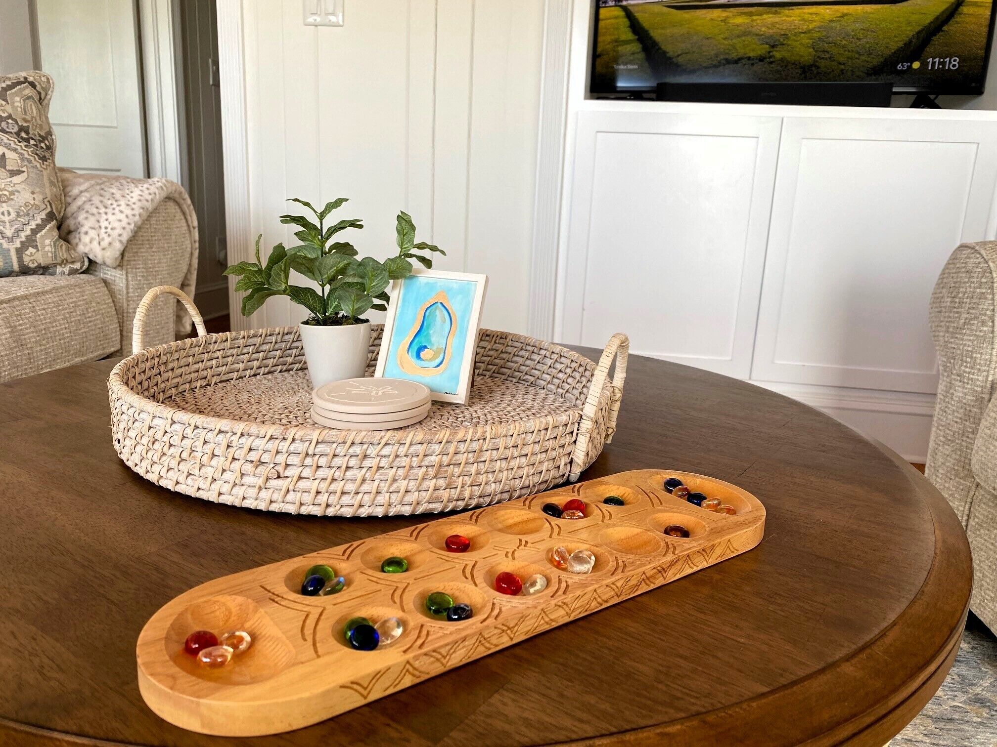 A round wooden table with a woven basket, a small potted plant, a framed picture, and a wooden tray with colorful marbles.