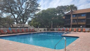 A rectangular swimming pool with a metal ladder, surrounded by red lounge chairs and a brick patio.