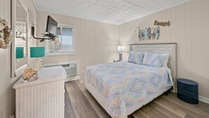 A bedroom with a bed, a nightstand, a lamp, a wall-mounted heater, a window with blinds, a wall decoration, and a small trash bin.