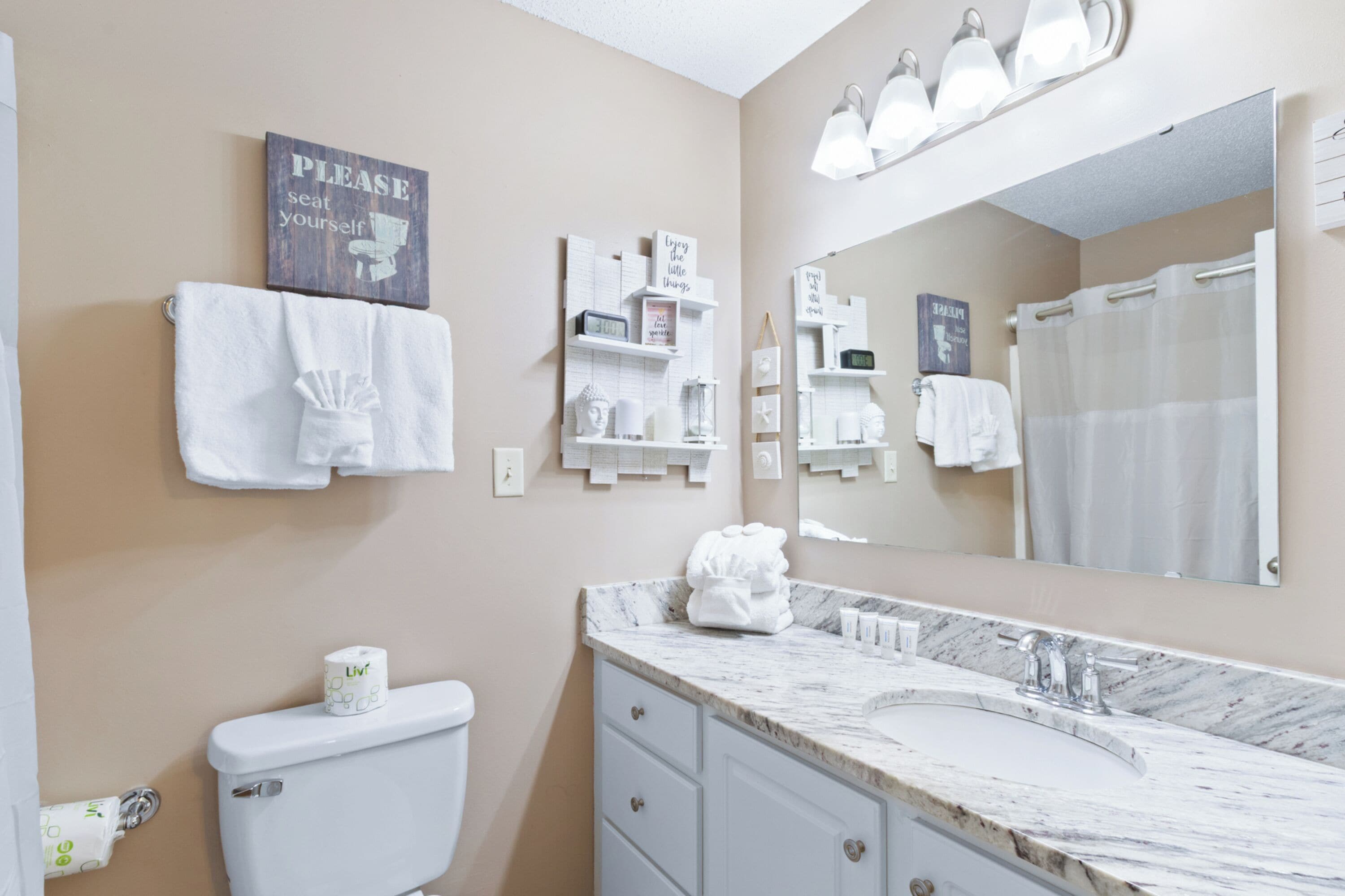 A bathroom with a toilet, sink, and mirror. There are towels hanging on the wall and a sign that says "PLEASE seat yourself."
