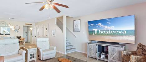 A spacious living room with a large flat-screen TV, comfortable seating, a ceiling fan, and a staircase leading to another level.