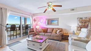 A living room with a sofa, armchairs, a coffee table, and a ceiling fan. There is a TV mounted on the wall and a window with a view of the outdoors. - Summer House West C201 (Gulf Shores)