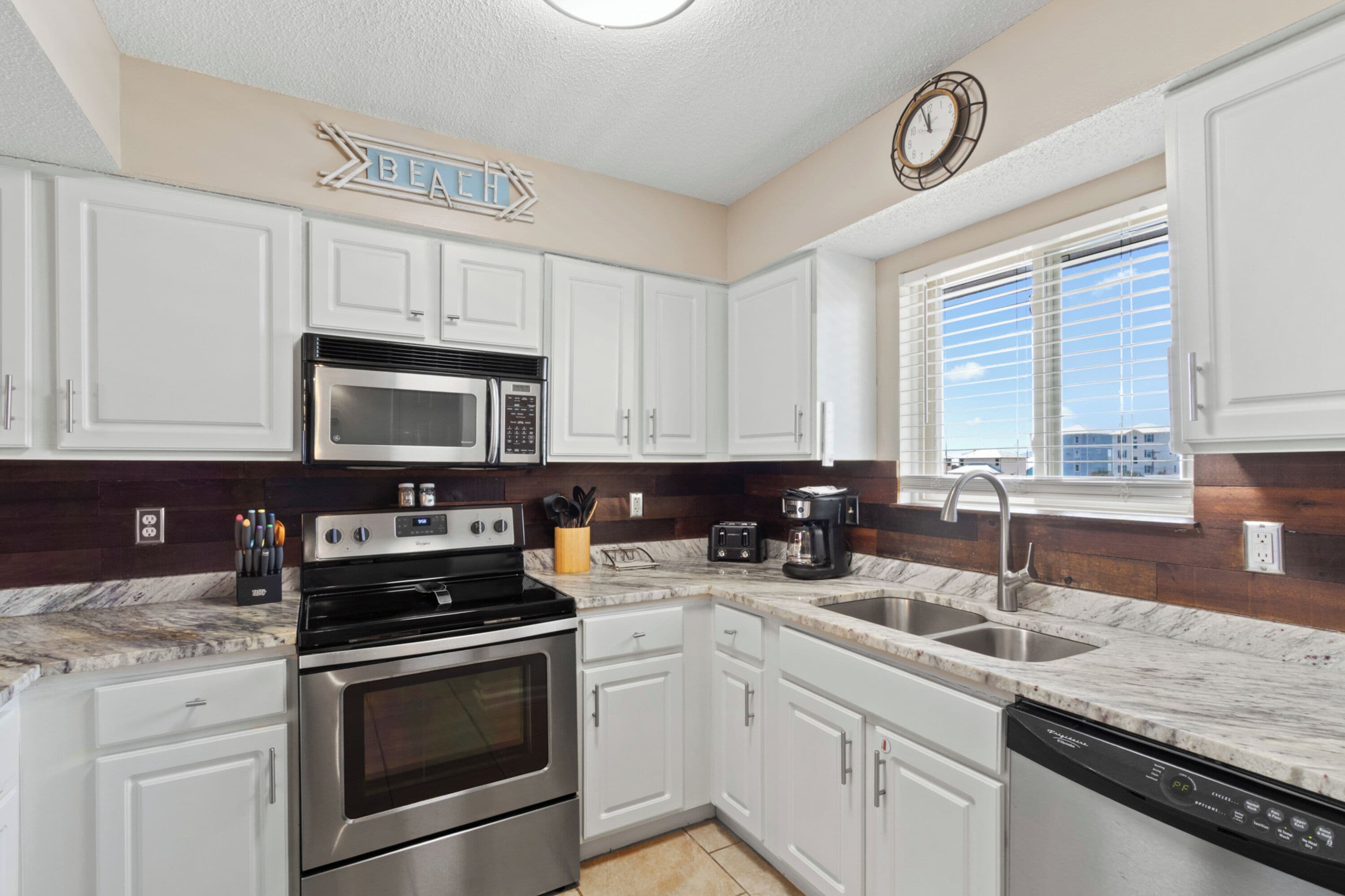 A modern kitchen with white cabinets, stainless steel appliances, a marble countertop, and a window with blinds.